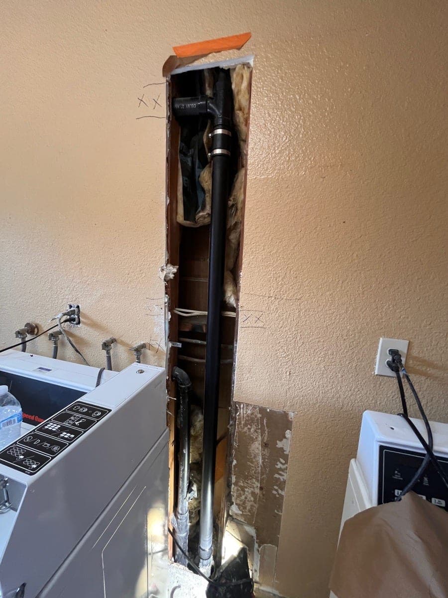 Before photo of a tall drywall opening exposing plumbing behind laundry equipment.